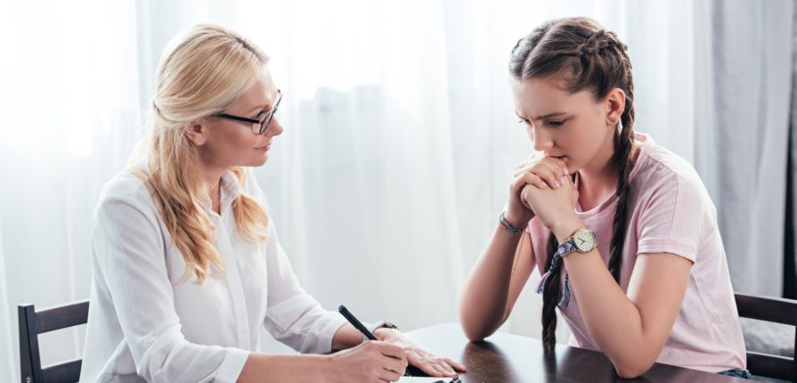 stressed teenage girl sitting at table on therapy session by female counselor writing in clipboard in office